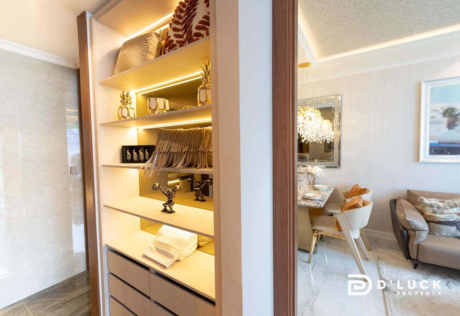 The-Riviera-Malibu-1bedroom 14 Open built-in wardrobe with illuminated shelves displaying gold pineapple decor, towels, and hanging items; dining area visible beyond.