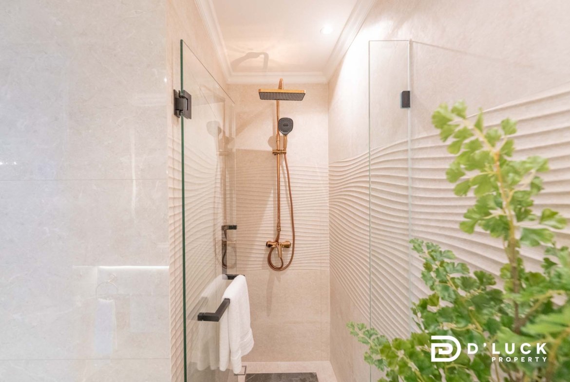 Modern glass-walled shower with a square rain showerhead and handheld hose; plant visible on the right.