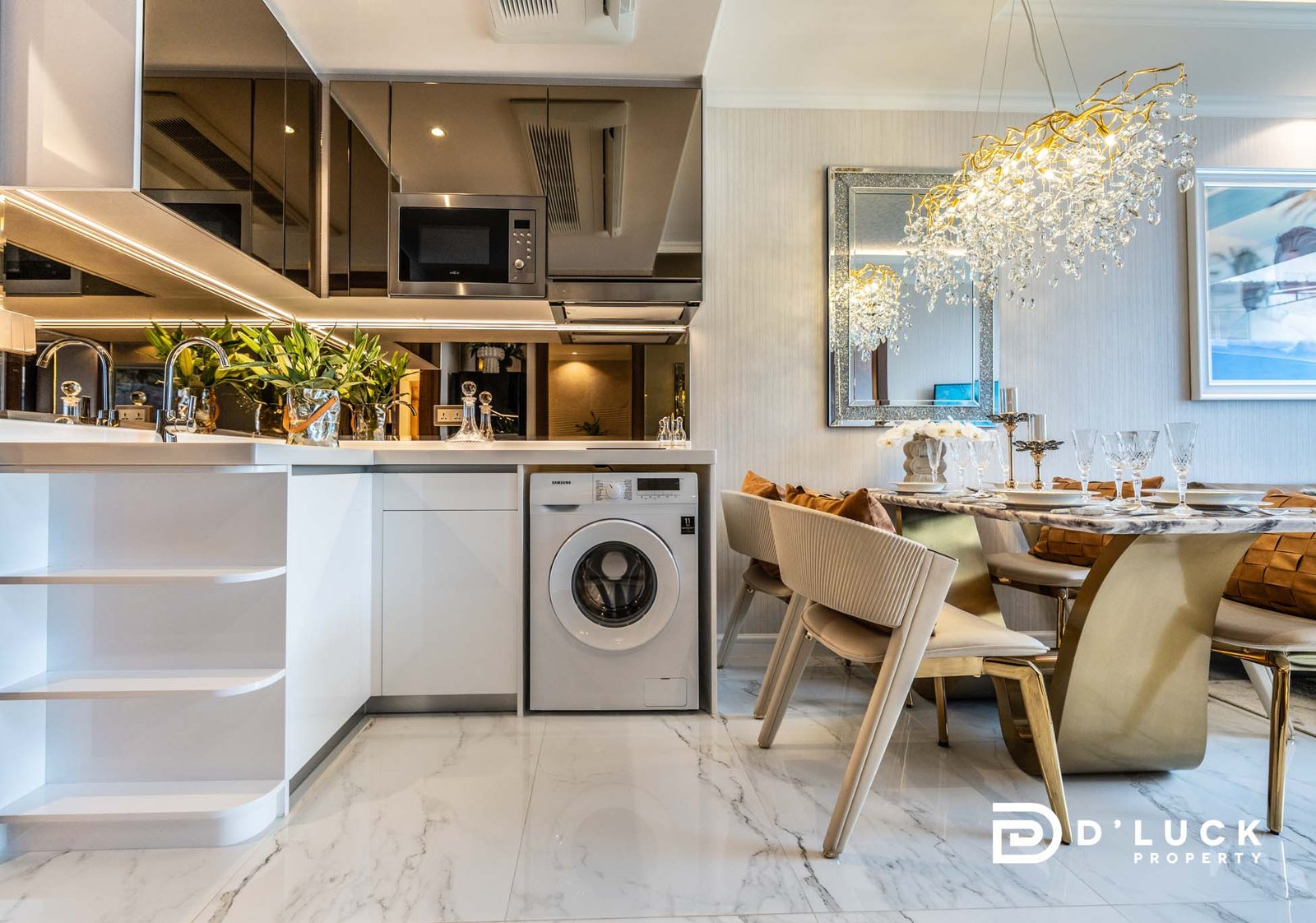 The-Riviera-Malibu-1bedroom 3 Modern open-plan kitchen with stainless appliances, marble floor, and a glass chandelier over a dining table set with gold accents. Logo bottom right says 'Lucky Property'.