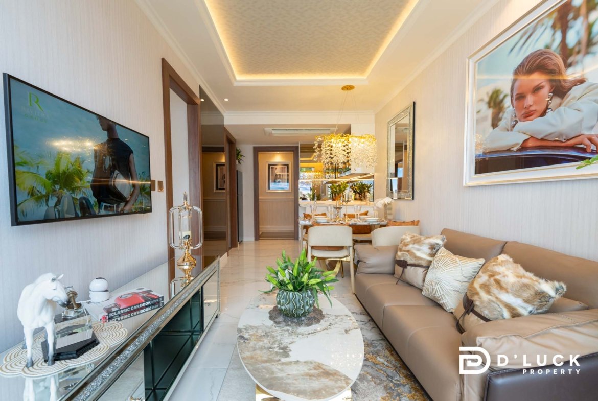 Luxurious modern living room with beige sectional, glass coffee tables, a wall-mounted TV on the left, and a large framed ad/photo on the right; gold chandelier and plants add elegance.