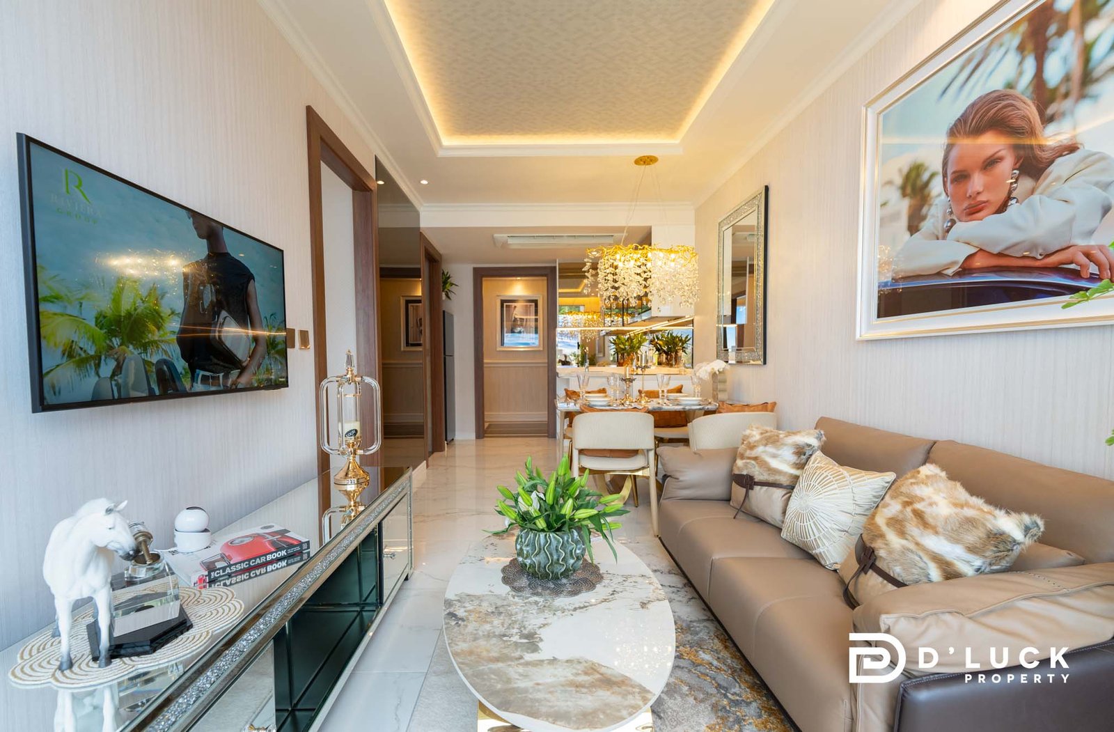 The-Riviera-Malibu-1bedroom 6 Luxurious modern living room with beige sectional, glass coffee tables, a wall-mounted TV on the left, and a large framed ad/photo on the right; gold chandelier and plants add elegance.