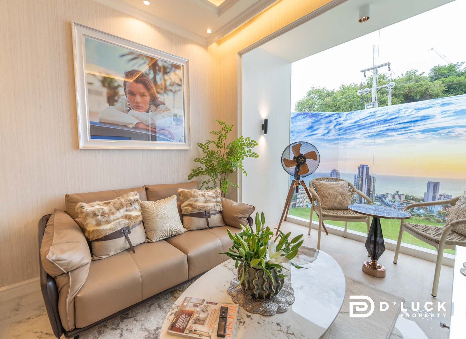 The-Riviera-Malibu-1bedroom 9 Modern living room with a beige leather sofa, fur cushions, and a glass coffee table; large window shows city skyline and ocean view.
