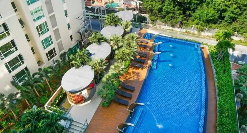 Aerial view of a hotel pool area with blue water, wooden deck, lounge chairs, palm trees, and a white building on the left.