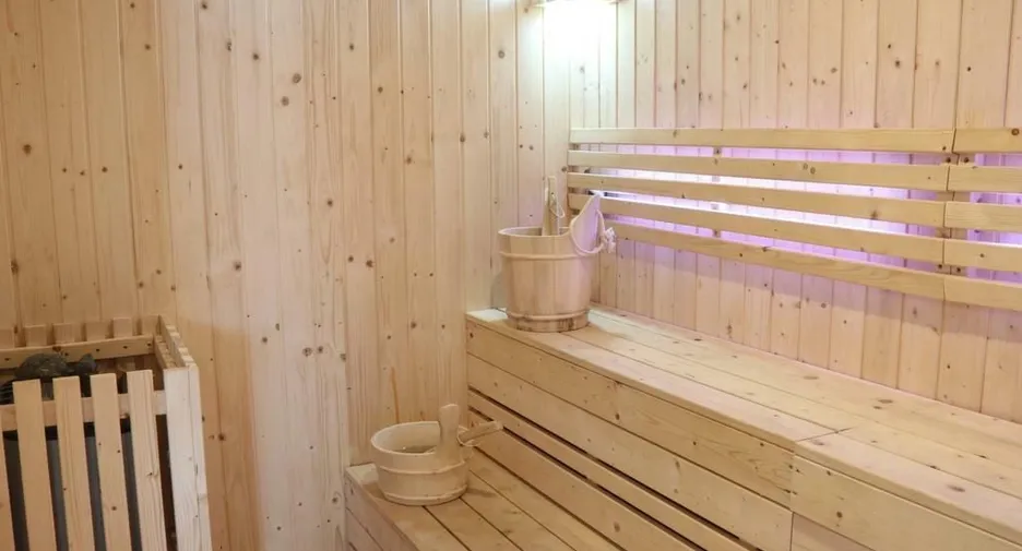 Wood-paneled sauna interior with long wooden benches, a bucket and ladle on the bench, and a heater with stones in the corner.