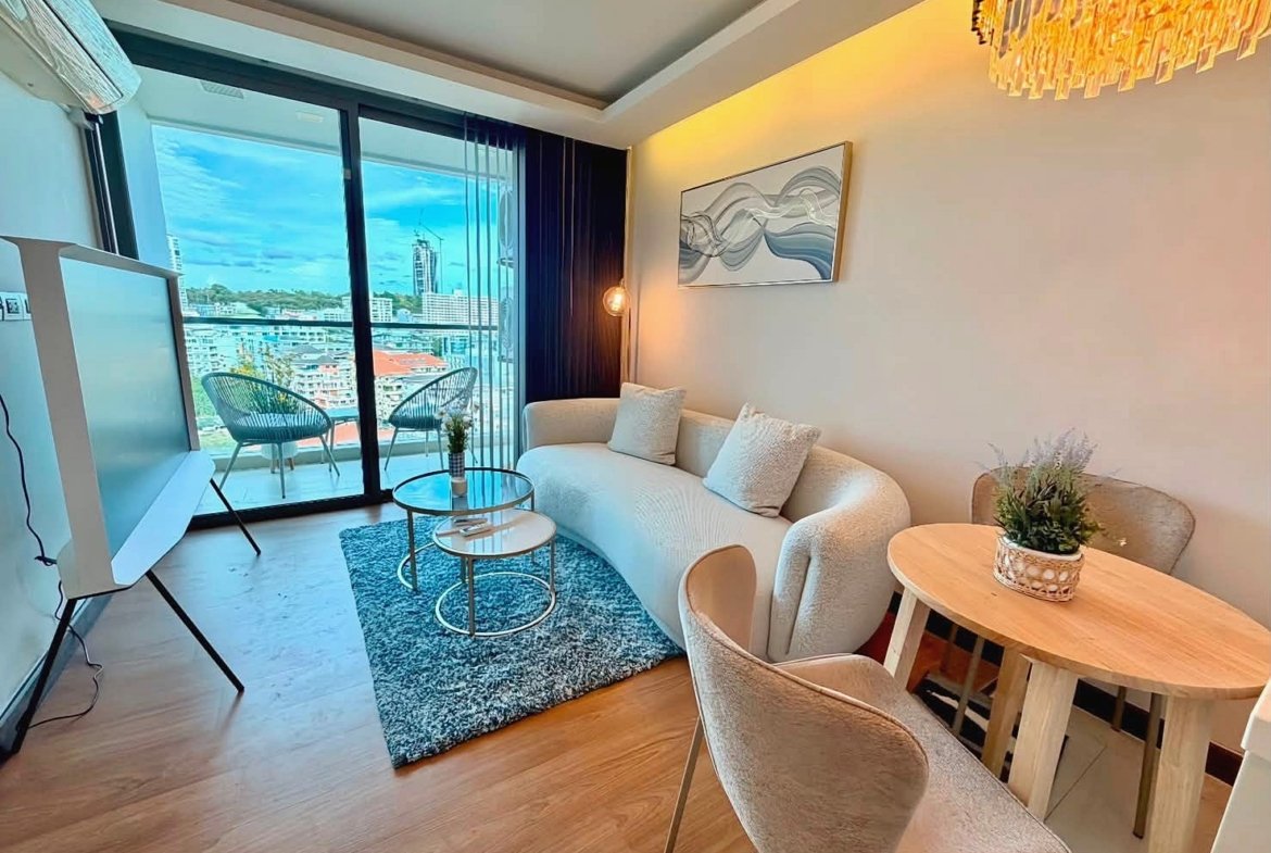 Modern living room with a curved cream sofa, two beige chairs, a glass coffee table, and a blue rug near sliding balcony doors showing a city view.