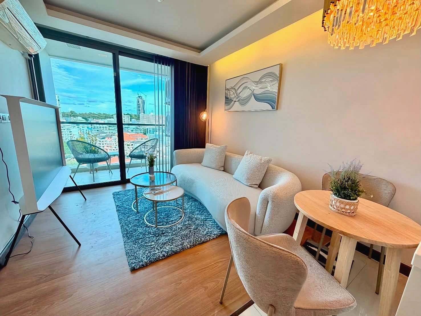 The_Peak_Tower_Rent_Rdl340 Modern living room with a curved cream sofa, two beige chairs, a glass coffee table, and a blue rug near sliding balcony doors showing a city view.