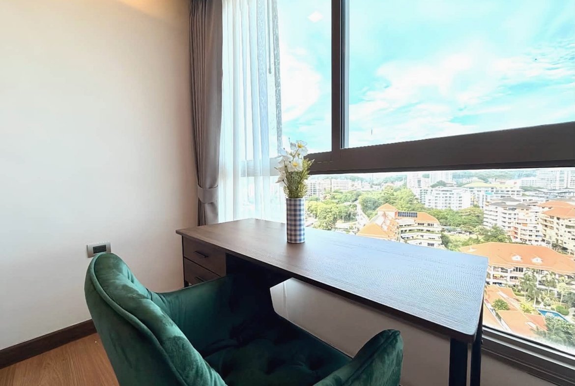 Home office corner with a dark wooden desk, green velvet chair, and a vase of flowers near a large window overlooking a cityscape.
