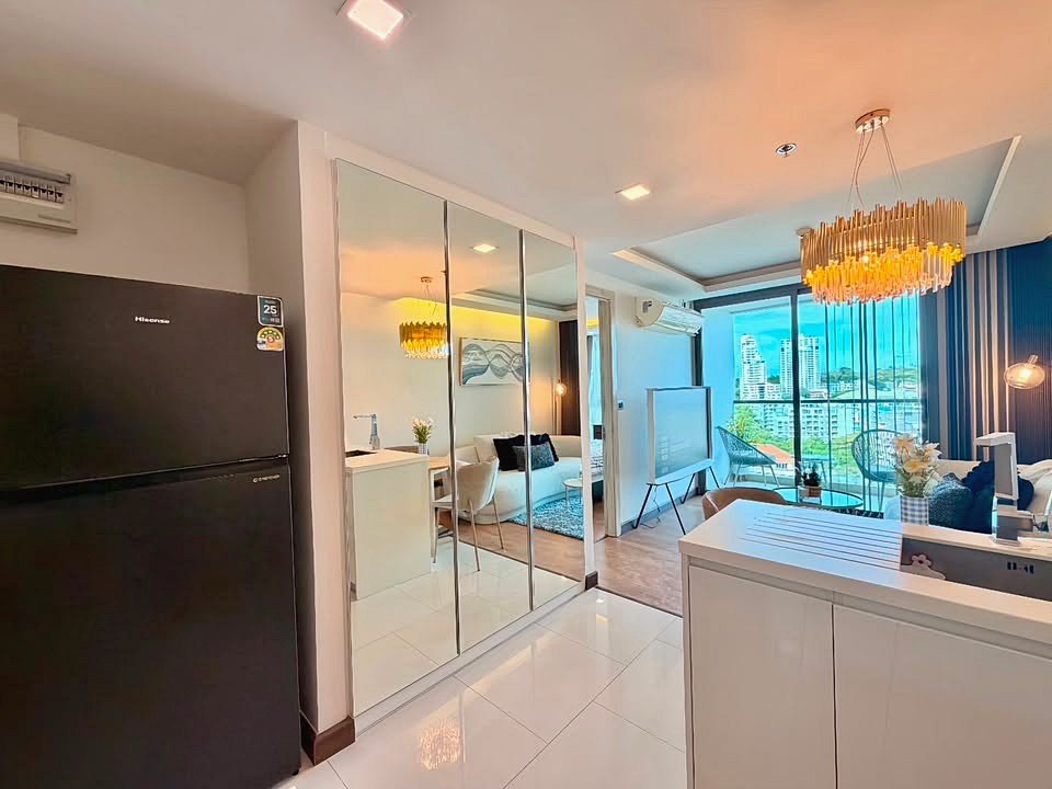 Open-plan living area with a black refrigerator, mirrored sliding wardrobe doors, and a chandelier over a seating area. The city skyline is visible through large windows in the background?