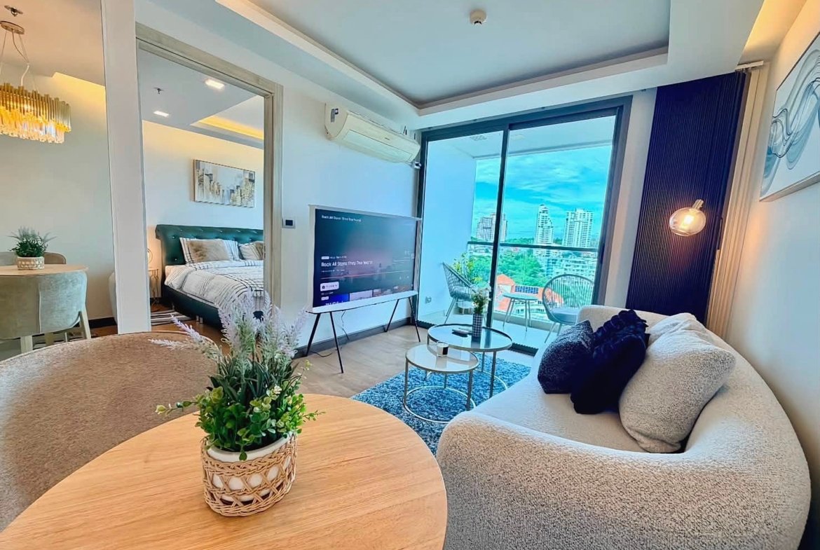 Open-plan living area with a beige curved sofa, round coffee tables, and a wall-mounted TV, overlooking a balcony with city views.
