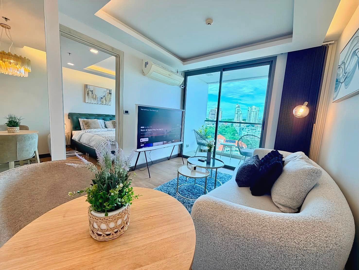 The_Peak_Tower_Rent_Rdl340_16 Open-plan living area with a beige curved sofa, round coffee tables, and a wall-mounted TV, overlooking a balcony with city views.