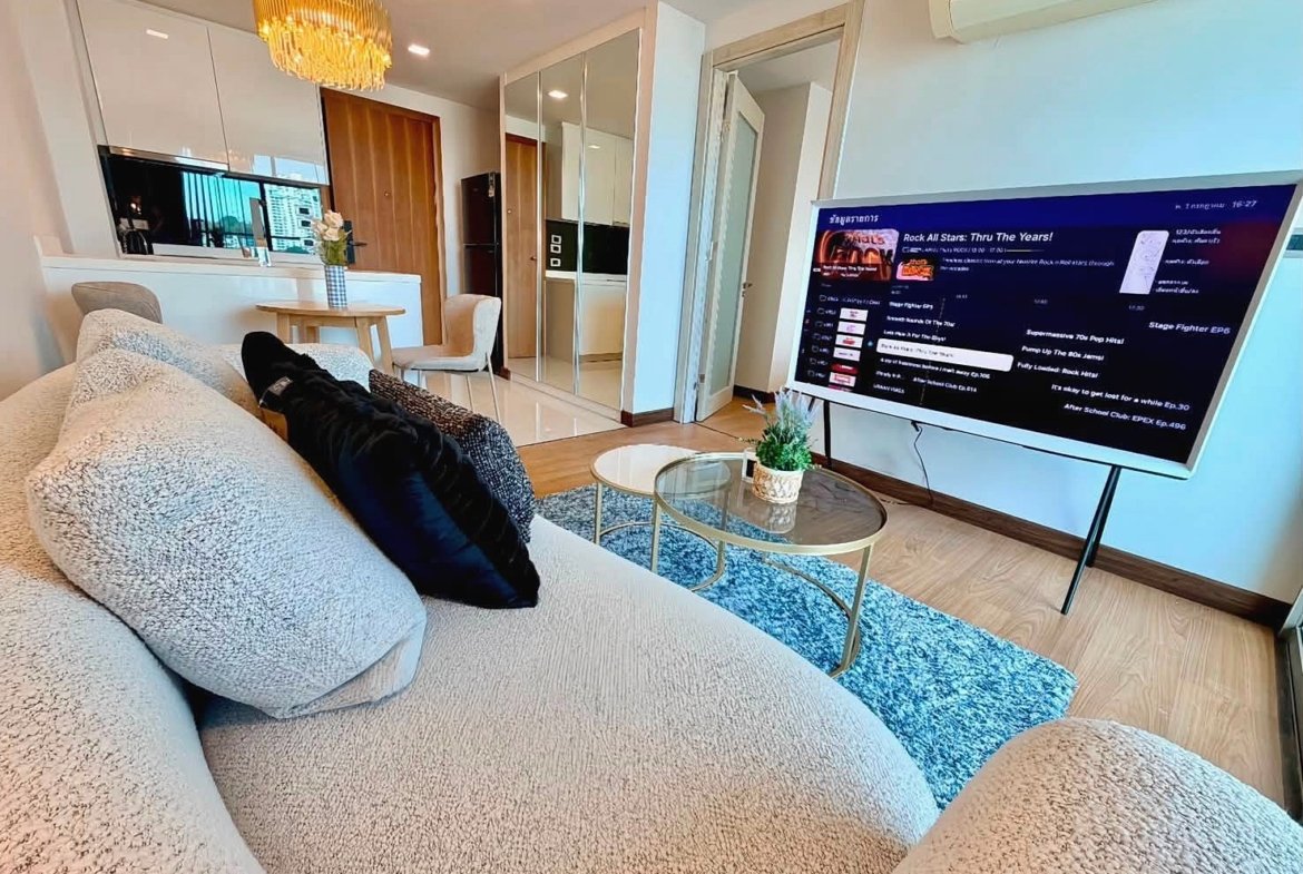 Cozy modern living room with a beige sectional sofa facing a large flat-screen TV, glass coffee table, and blue rug; open kitchen in the background.
