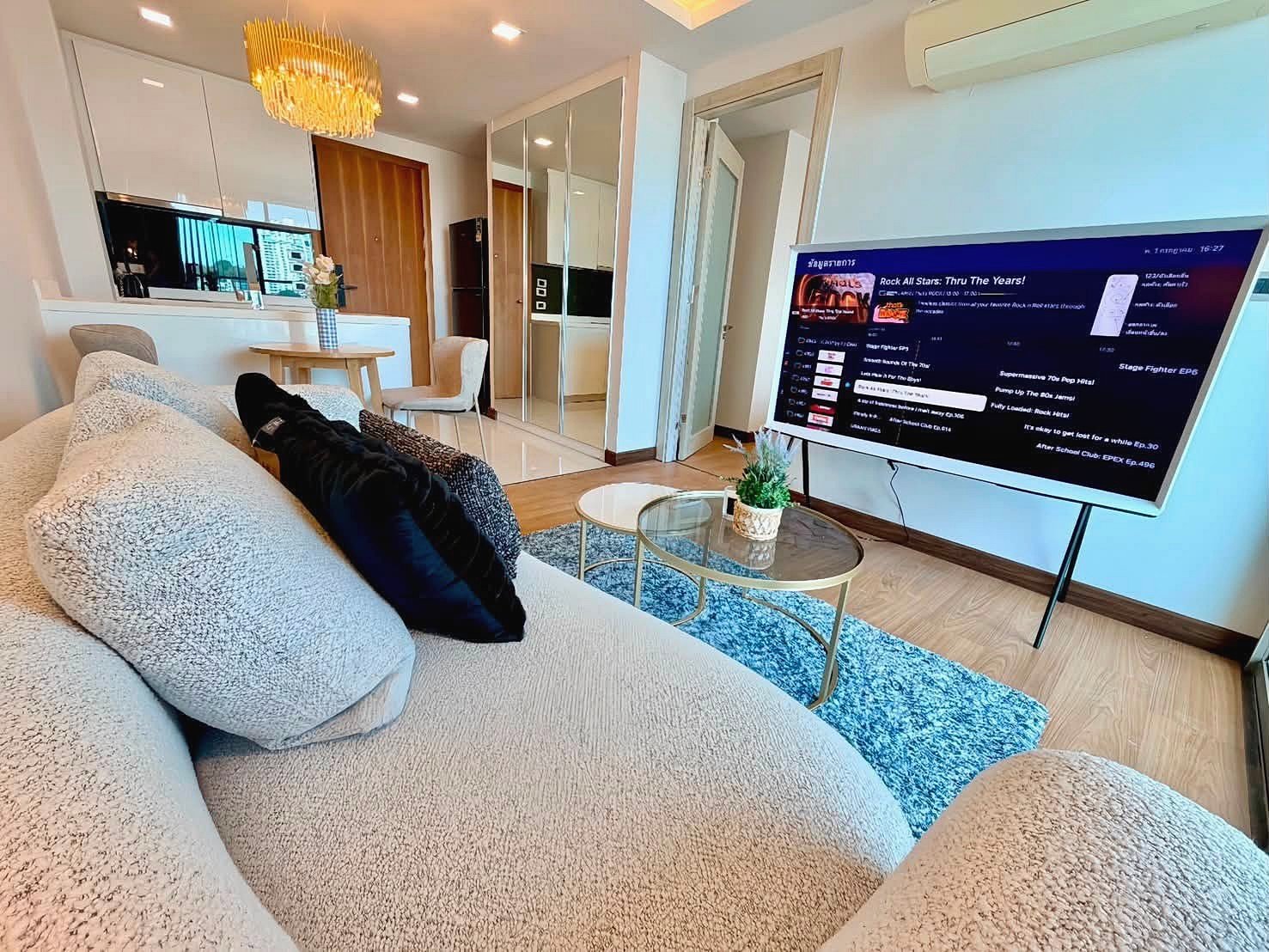 The_Peak_Tower_Rent_Rdl340_2 Cozy modern living room with a beige sectional sofa facing a large flat-screen TV, glass coffee table, and blue rug; open kitchen in the background.