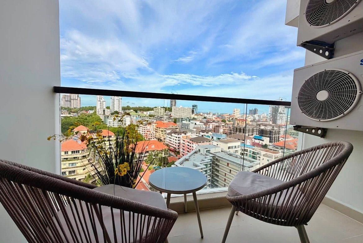Balcony seating area with two woven chairs and a small round table, overlooking a city skyline on a sunny day.