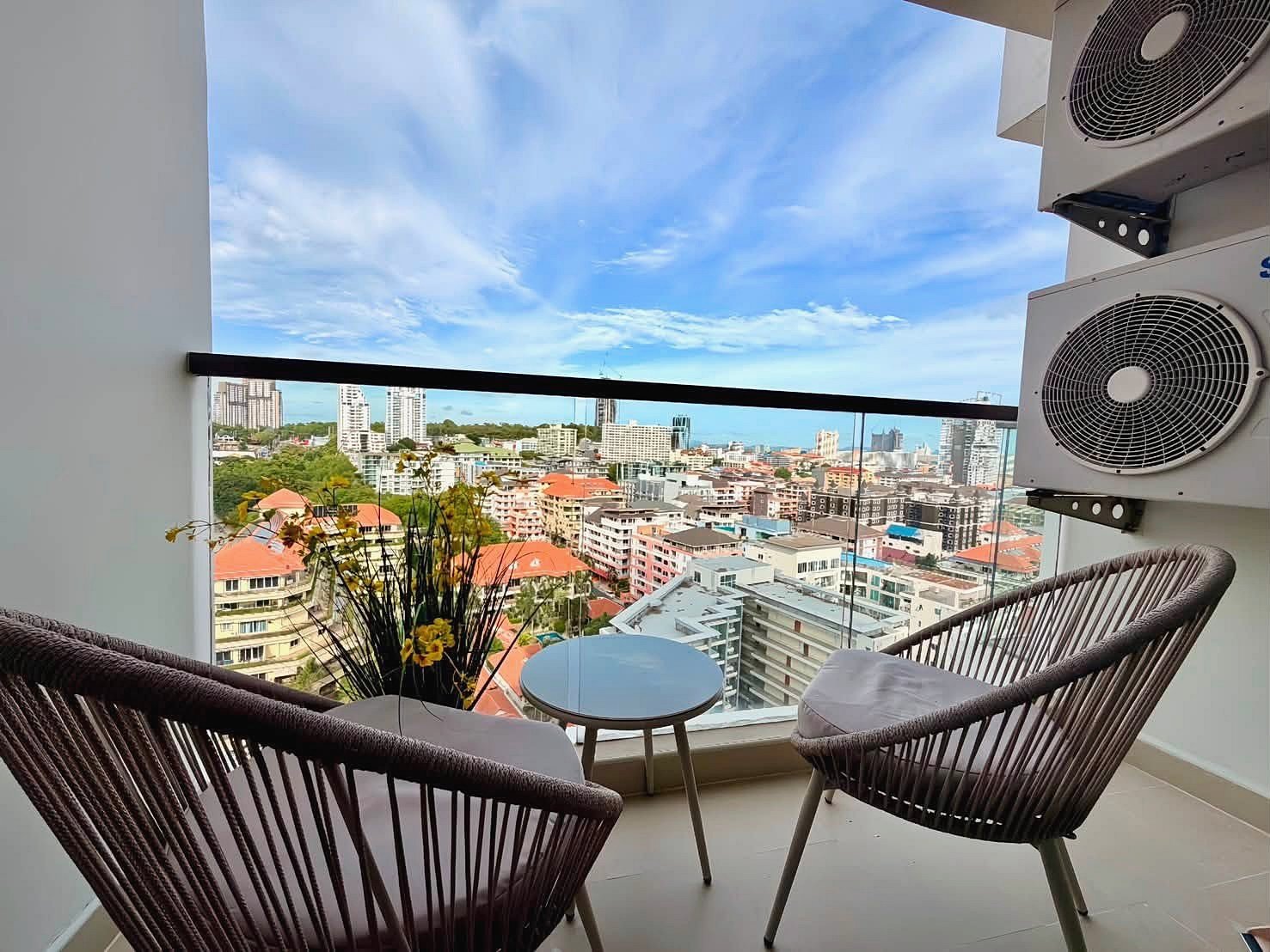 The_Peak_Tower_Rent_Rdl340_21 Balcony seating area with two woven chairs and a small round table, overlooking a city skyline on a sunny day.