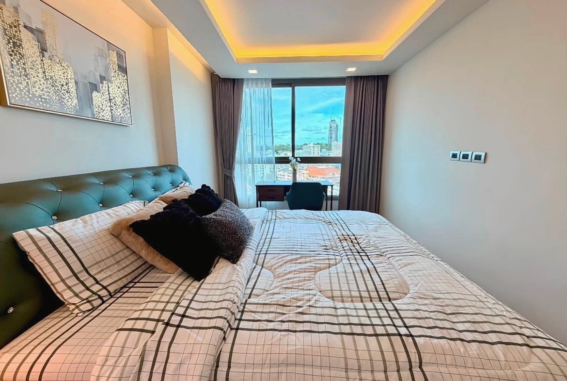 Modern hotel bedroom with a green tufted headboard, plaid bedding, and decorative pillows, plus a desk by the window with a city view.