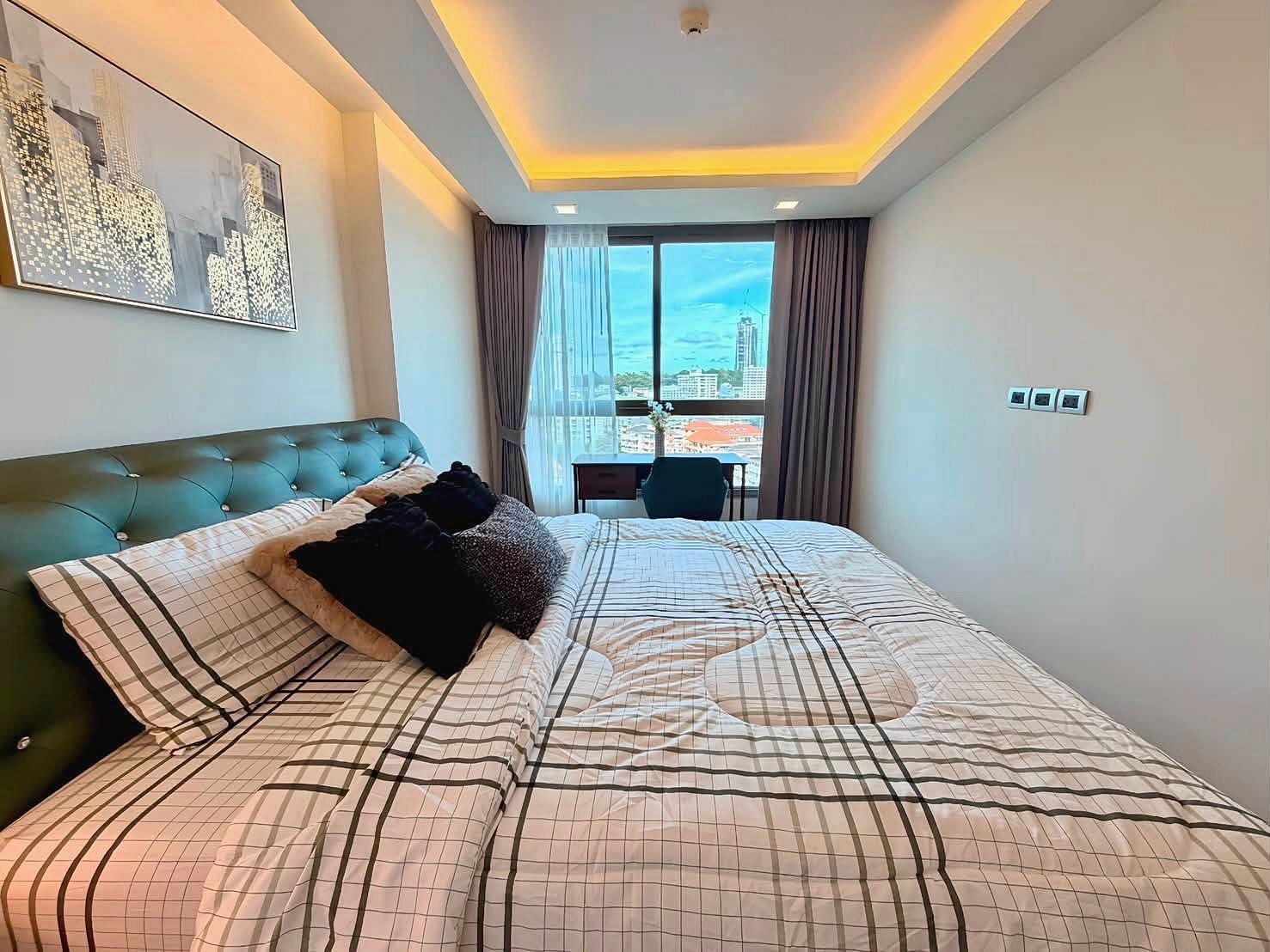 The_Peak_Tower_Rent_Rdl340_6 Modern hotel bedroom with a green tufted headboard, plaid bedding, and decorative pillows, plus a desk by the window with a city view.