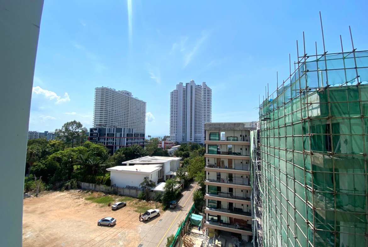 Construction site with scaffolding and green safety netting on a tall building against a bright blue sky and surrounding low-rise blocks left and trees below.