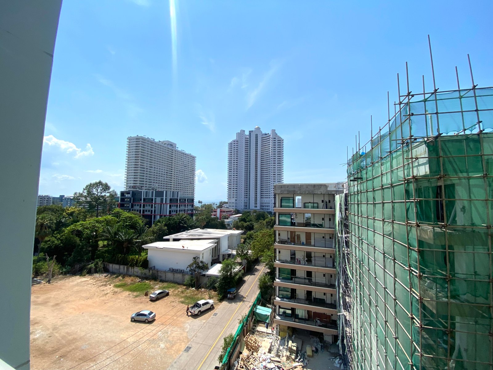 Construction site with scaffolding and green safety netting on a tall building against a bright blue sky and surrounding low-rise blocks left and trees below.