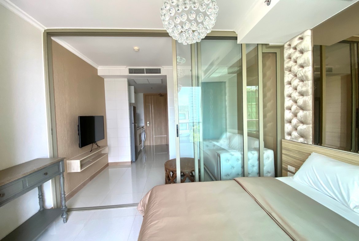 Modern hotel bedroom with a beige bedspread, glass sliding doors, and a crystal chandelier overhead.
