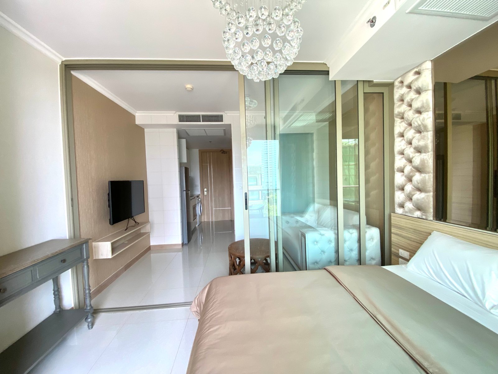 Modern hotel bedroom with a beige bedspread, glass sliding doors, and a crystal chandelier overhead.