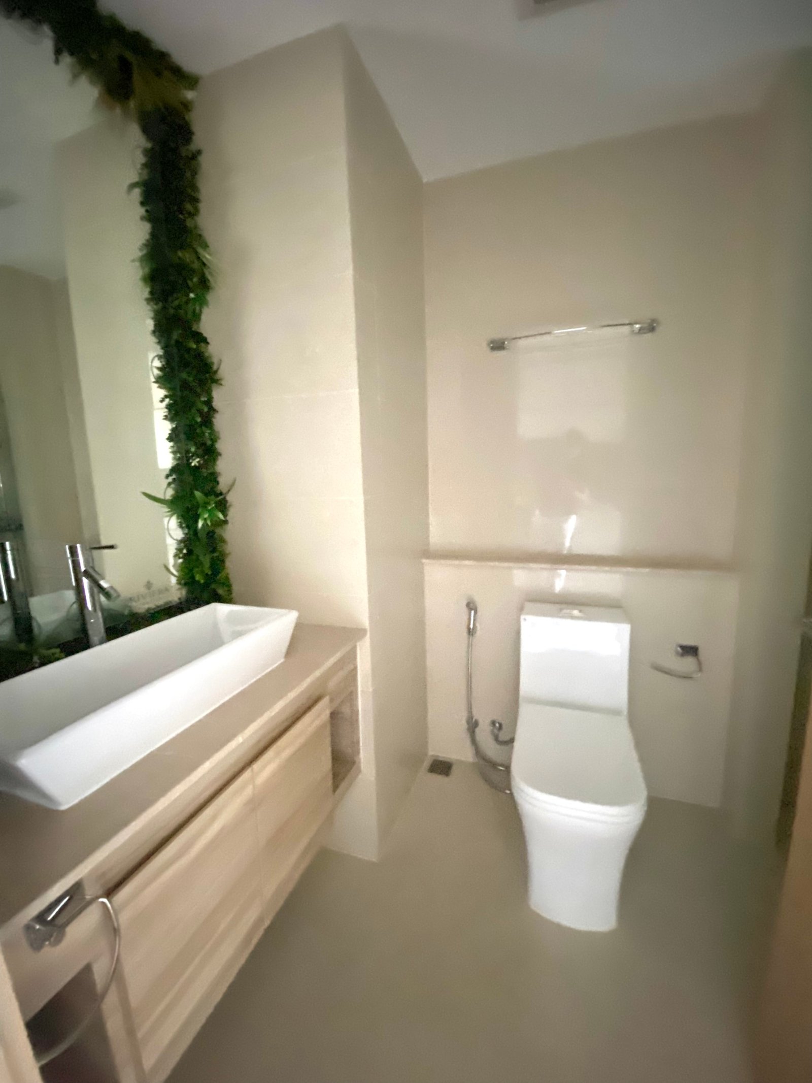 Modern bathroom with a white rectangular sink atop a wooden vanity and a wall mirror; greenery runs along the wall behind the sink.