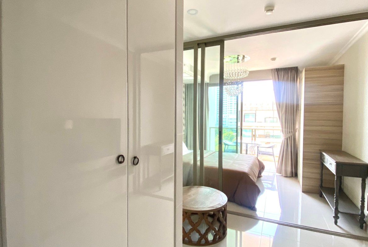 Bright modern hotel bedroom with white wardrobe, glass partition, and a bed by a sunlit balcony.