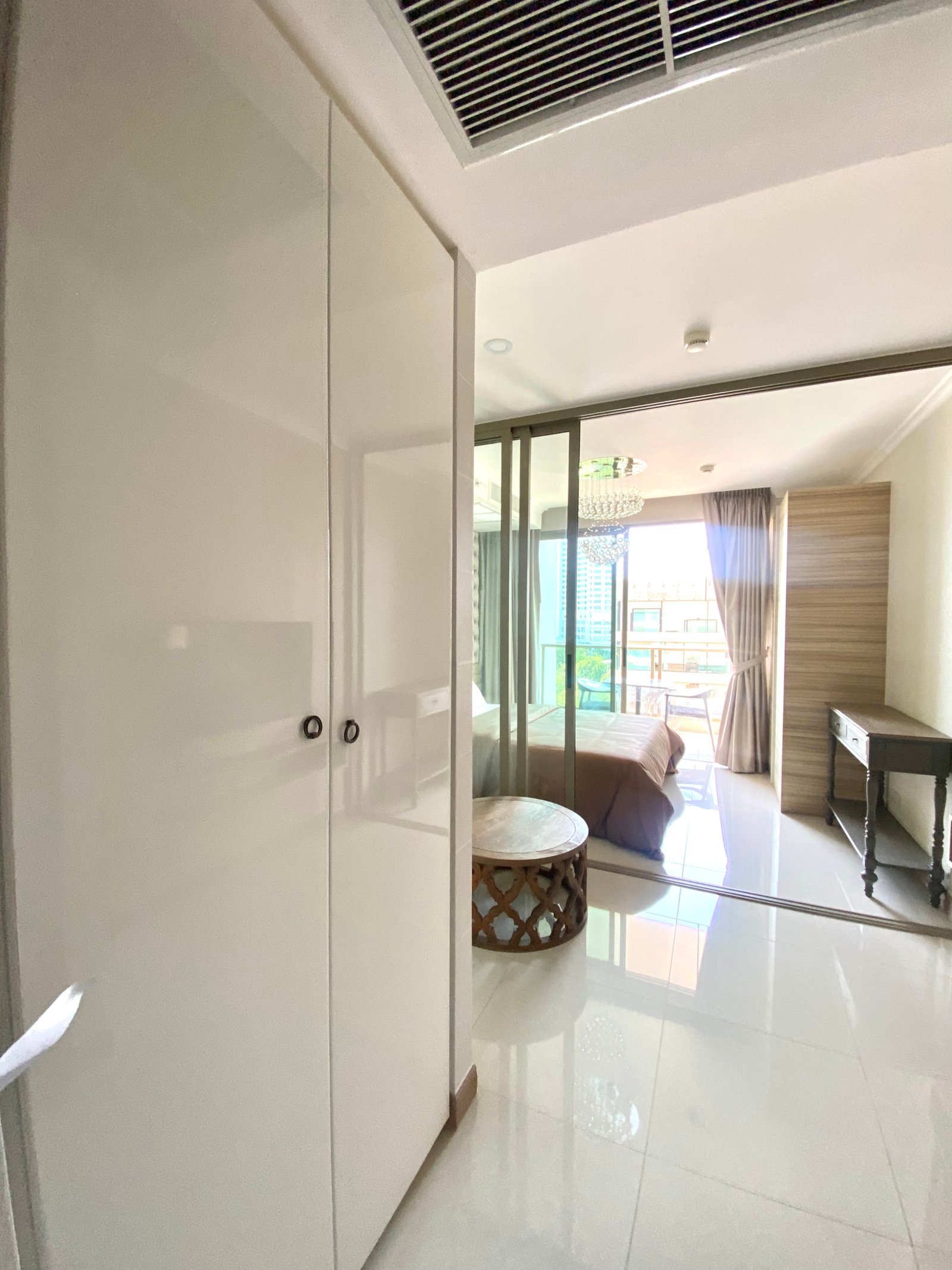 Bright modern hotel bedroom with white wardrobe, glass partition, and a bed by a sunlit balcony.