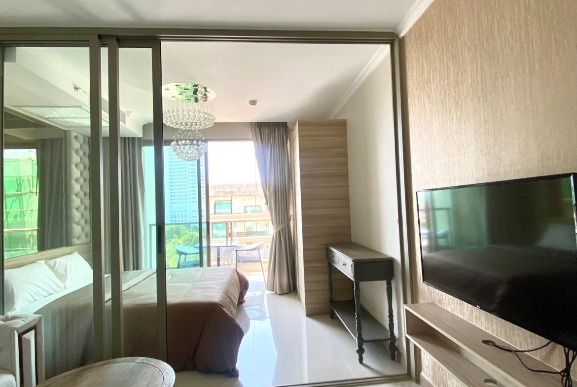 Hotel room with glass partition, two beds on the left, and a balcony with outdoor seating beyond; chandelier visible in the adjacent living area.