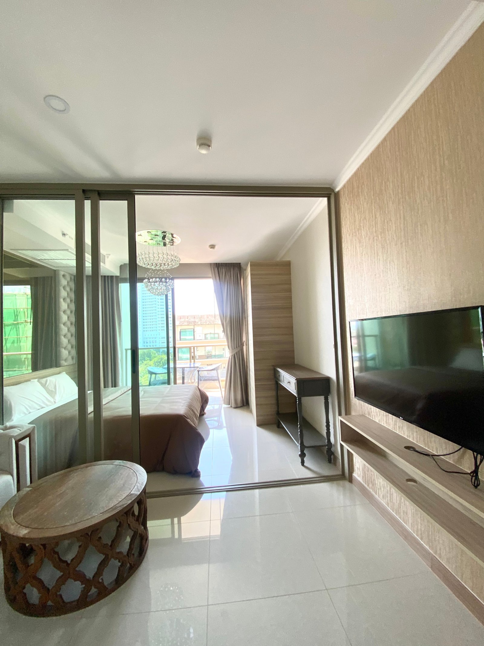 Hotel room with glass partition, two beds on the left, and a balcony with outdoor seating beyond; chandelier visible in the adjacent living area.