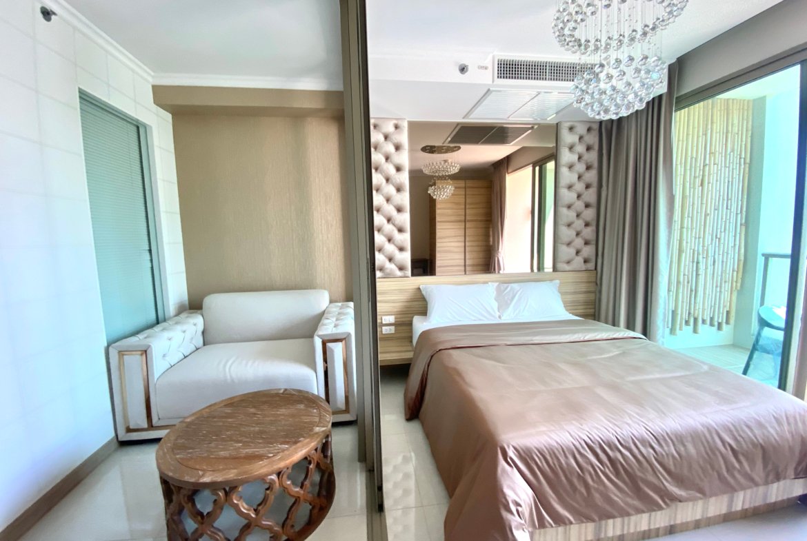 Hotel bedroom with twin beige beds, tufted headboard, and a round carved wooden table; chandelier overhead. (View through a sliding glass door)