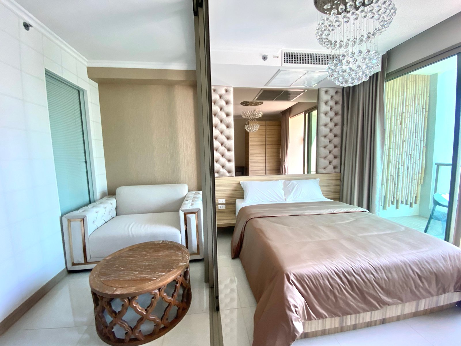 Hotel bedroom with twin beige beds, tufted headboard, and a round carved wooden table; chandelier overhead. (View through a sliding glass door)