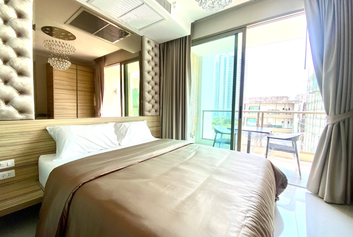 Hotel bedroom with a large tufted headboard, two twin beds, and wooden wall panels; floor-to-ceiling windows with sheer curtains and a balcony visible outside