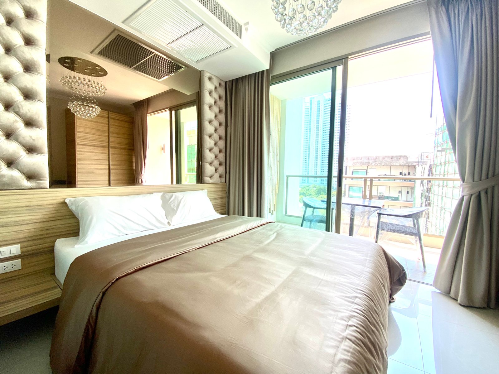 Hotel bedroom with a large tufted headboard, two twin beds, and wooden wall panels; floor-to-ceiling windows with sheer curtains and a balcony visible outside