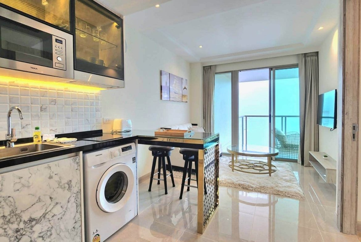 Modern open-plan kitchen with marble countertops, stainless steel appliances, washing machine, and a bar-height island with two stools; balcony and ocean view beyond.