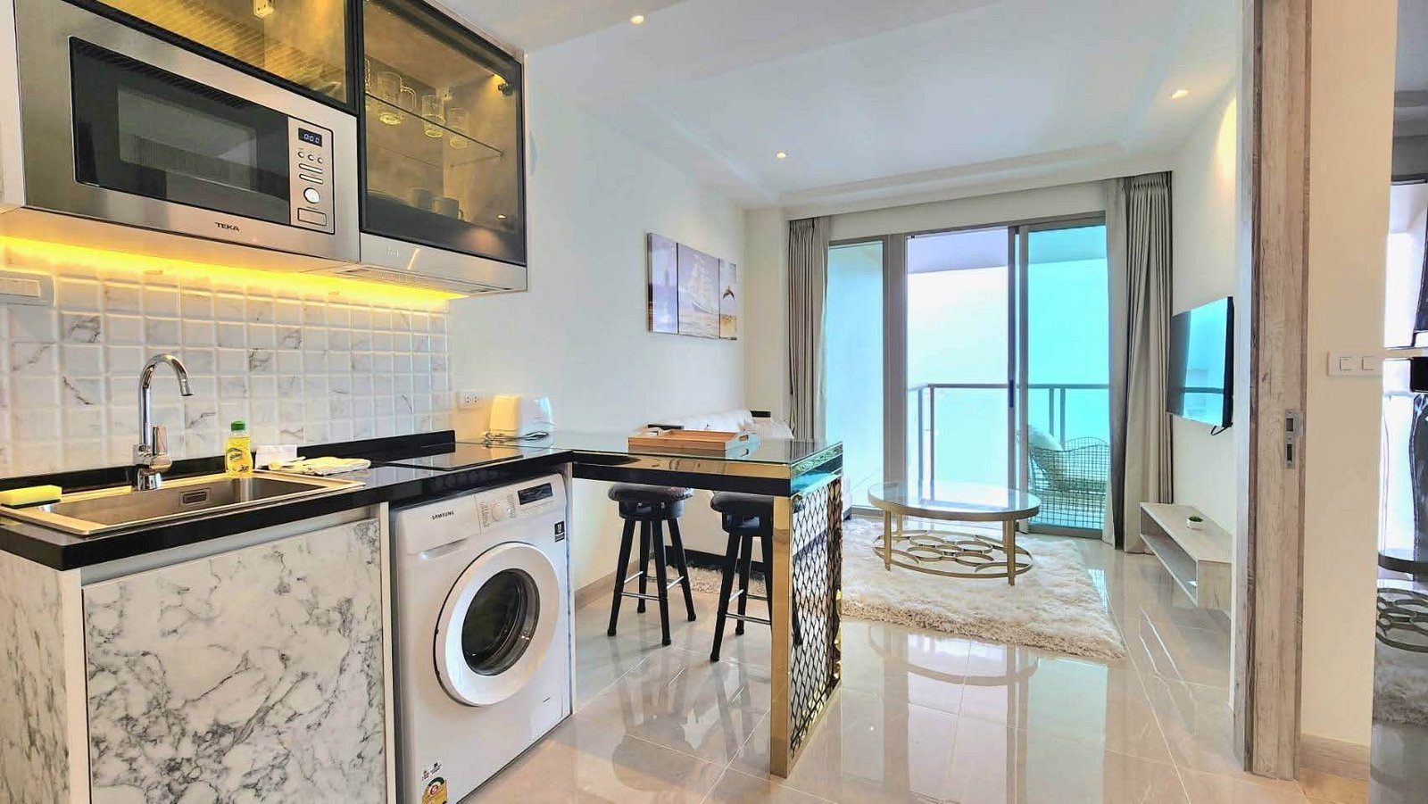 Modern open-plan kitchen with marble countertops, stainless steel appliances, washing machine, and a bar-height island with two stools; balcony and ocean view beyond.