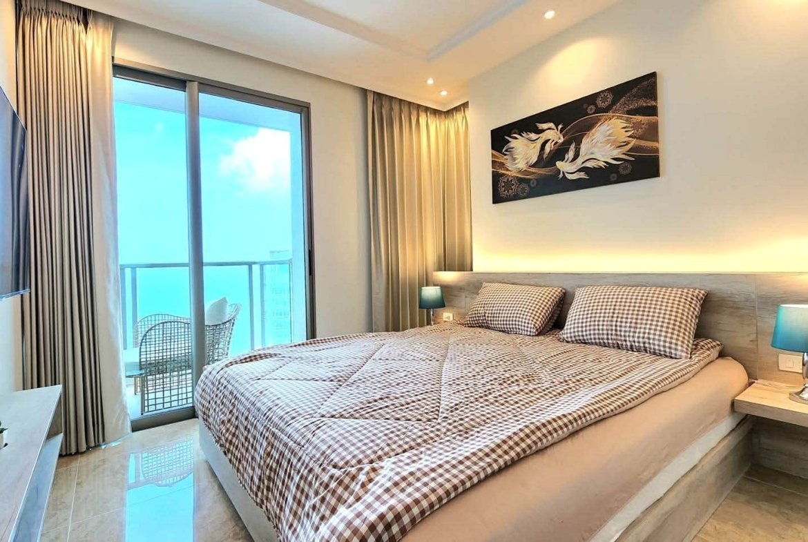 Hotel bedroom with a king bed, checkered duvet, and two teal bedside lamps; balcony doors open to a seating area outside.