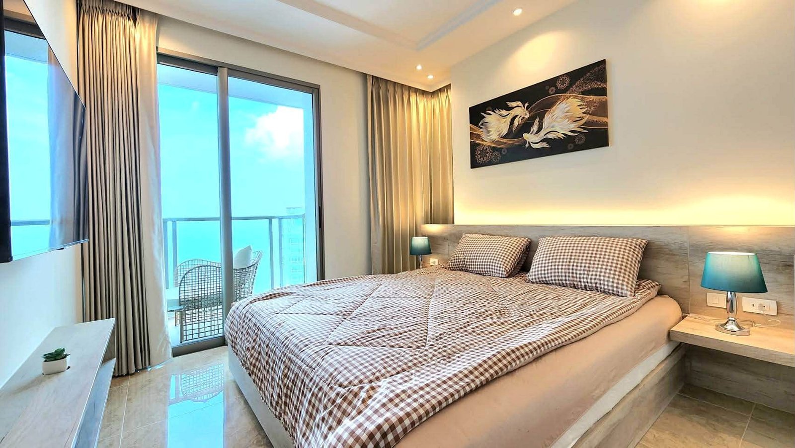 Hotel bedroom with a king bed, checkered duvet, and two teal bedside lamps; balcony doors open to a seating area outside.