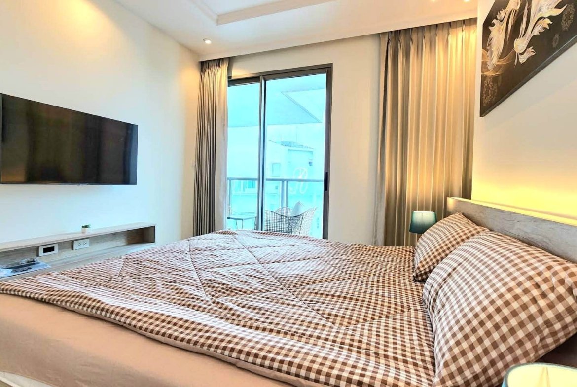 Hotel bedroom with a large bed, brown-and-white checkered bedding, and a tufted headboard, soft warm lighting along the wall, and a mounted flat-screen TV across from the bed.