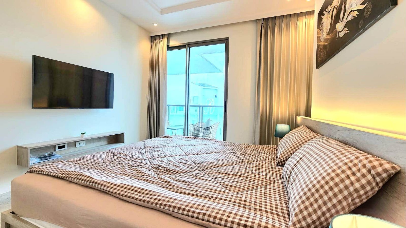Hotel bedroom with a large bed, brown-and-white checkered bedding, and a tufted headboard, soft warm lighting along the wall, and a mounted flat-screen TV across from the bed.
