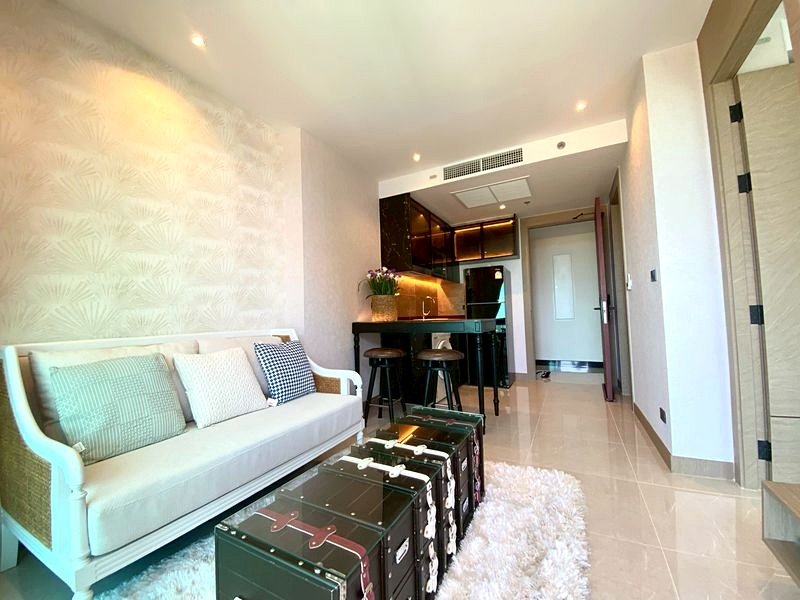 Bright studio living area with a white sofa and pastel cushions, stacked vintage trunk-style suitcases on a fluffy rug, open kitchen in the background.