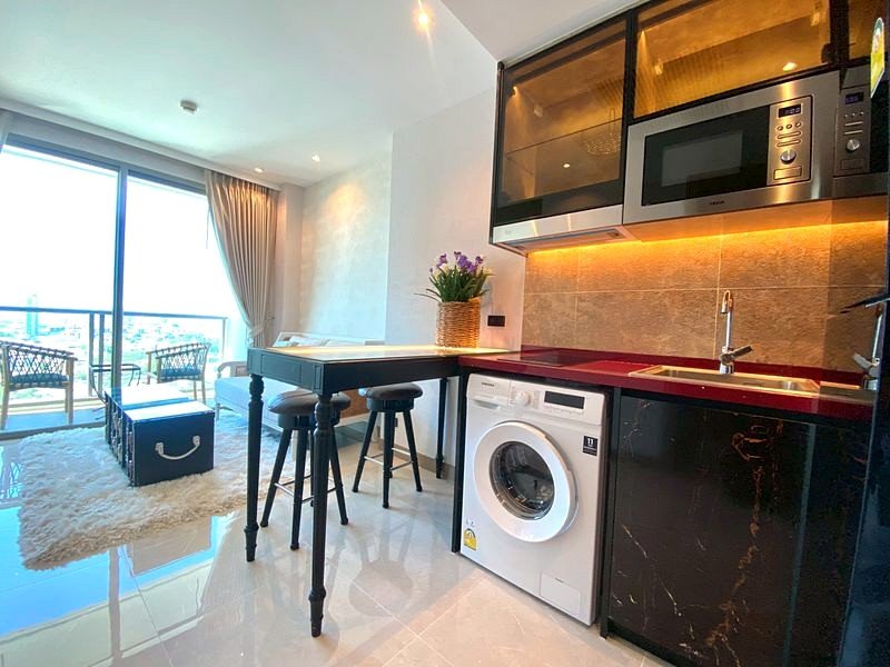 Modern open-plan kitchen with washing machine under red countertop, black cabinets, and stainless steel appliances, plus a balcony view in the background.
