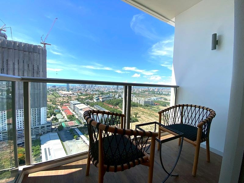 Balcony with two woven chairs and a small glass table, overlooking a cityscape beneath a blue sky.