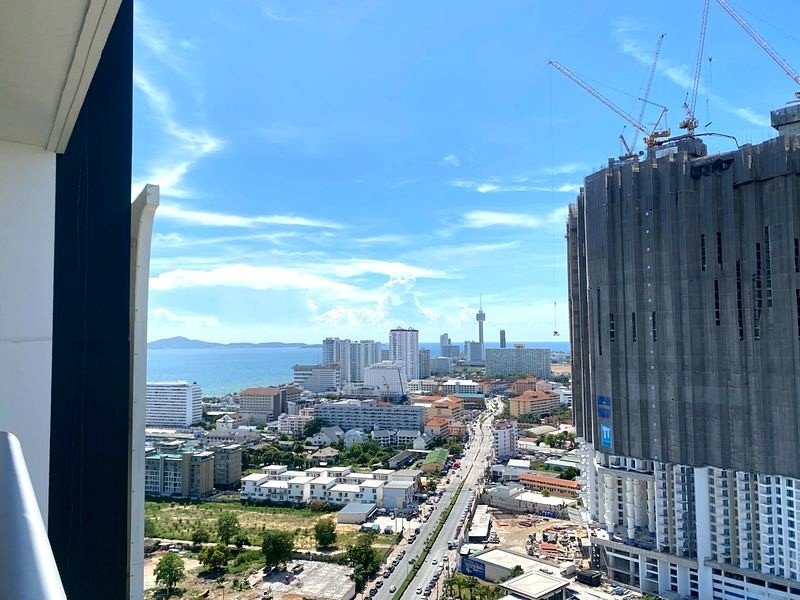 Coastal city skyline with a tall unfinished building and cranes on the right, blue ocean visible to the left. This high-rise view includes a road and mid-rise buildings toward the horizon.