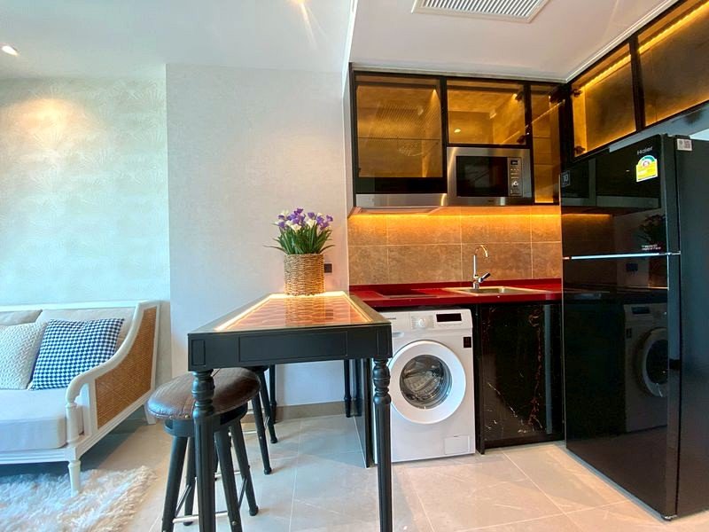 Open-plan kitchenette and living area with black cabinets, sink, washing machine, and fridge; a small table with a potted plant and two stools beside a white sofa.