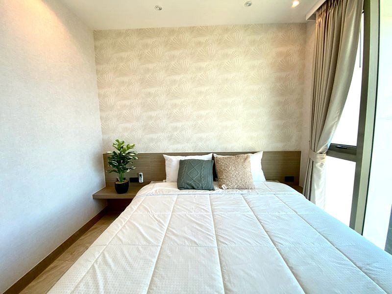 Modern hotel-style bedroom with a made bed, white quilt, decorative pillows, and a small plant on the floating nightstand by a patterned wallpapered wall.