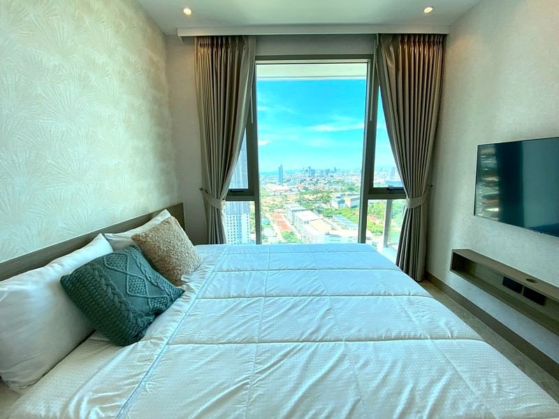 Hotel bedroom with a large window showing a city view, and a neatly made bed with white linens and decorative pillows.