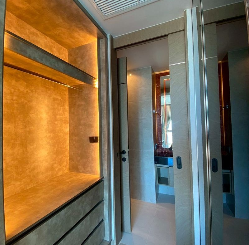 Warmly lit built-in cabinet with open storage and drawers along a beige wall, in a modern bathroom/closet area.