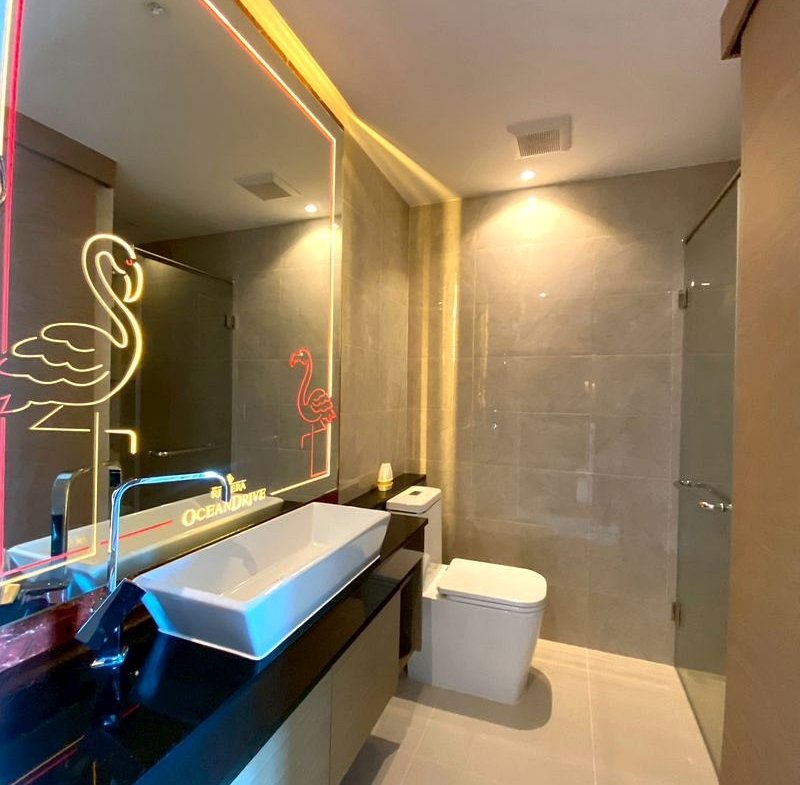 Modern bathroom with a white rectangular vessel sink, black countertop, and neon pink flamingo drawing on the backlit mirror beside a glass shower enclosure.