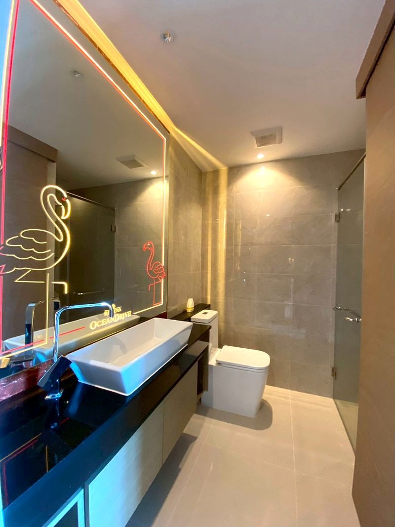 The_Riviera_Ocean_Drive_3014_8 Modern bathroom with a white rectangular vessel sink, black countertop, and neon pink flamingo drawing on the backlit mirror beside a glass shower enclosure.