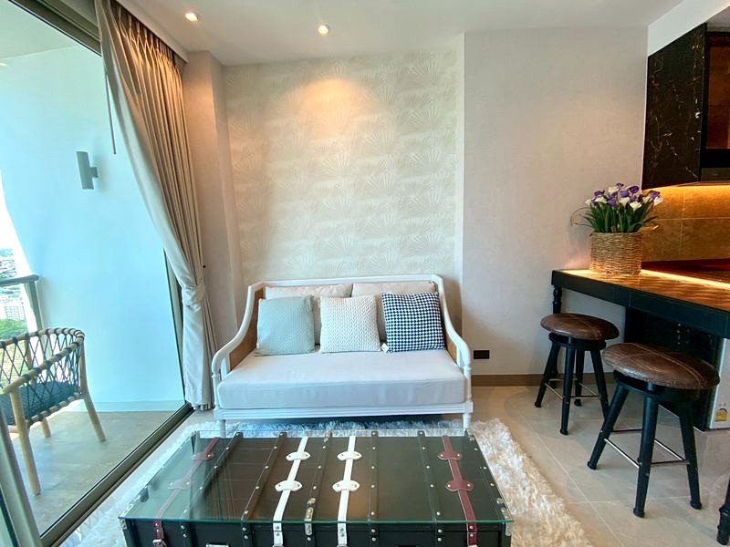 Bright living area with a white sofa and light cushions, a glass-trunk coffee table, and a balcony to the left showing city views.